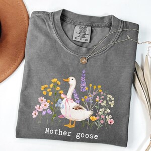 May include: Gray Comfort Colors t-shirt featuring a watercolor illustration of a goose with a pink bow, surrounded by colorful flowers. The text "Mother goose" is printed below the image. A gold pendant necklace and a brown hat are also visible.