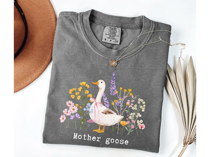 May include: Gray Comfort Colors t-shirt featuring a watercolor illustration of a goose with a pink bow, surrounded by colorful flowers. The text "Mother goose" is printed below the image. A gold pendant necklace and a brown hat are also visible.