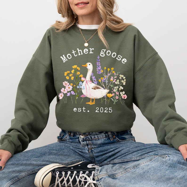 May include: Olive green sweatshirt with the words "Mother Goose" and "est. 2025" printed in white. A white goose with a pink bow is surrounded by colorful flowers. The sweatshirt is worn by a person.