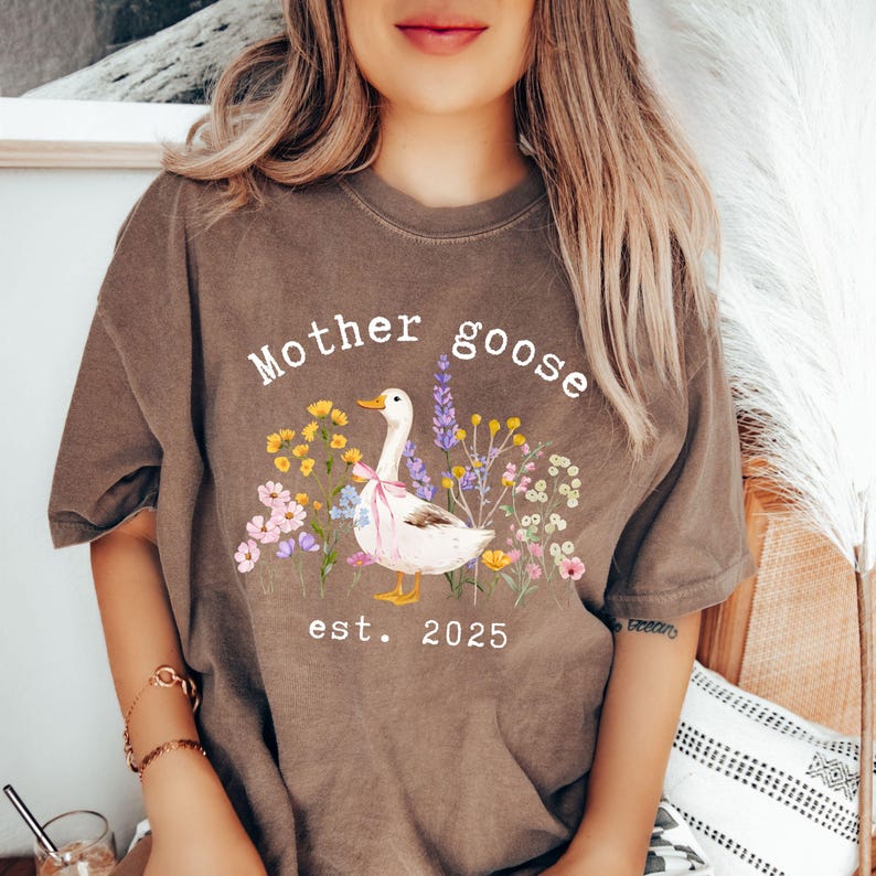 May include: Brown t-shirt with the text "Mother goose" above a graphic of a white goose with a pink bow, surrounded by colorful flowers. The text "est. 2025" is below the goose.