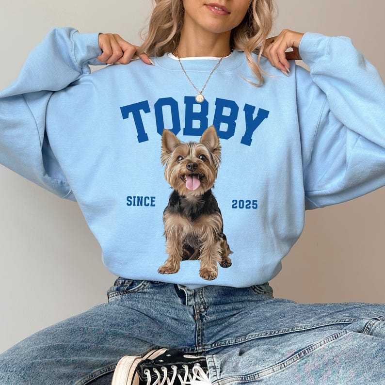 May include: Pale blue sweatshirt featuring the name "TOBBY" in navy blue block letters and a Yorkshire Terrier graphic. The text "SINCE 2025" is below the dog. The sweatshirt is worn by a person.