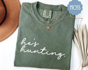 May include: A moss green t-shirt with the text "he's hunting." in white lettering.