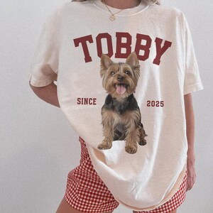 May include: Cream-coloured t-shirt featuring the name "TOBBY" in maroon block letters and a Yorkshire Terrier graphic. The text "SINCE 2025" is below the dog. The person is wearing red and white checkered shorts.