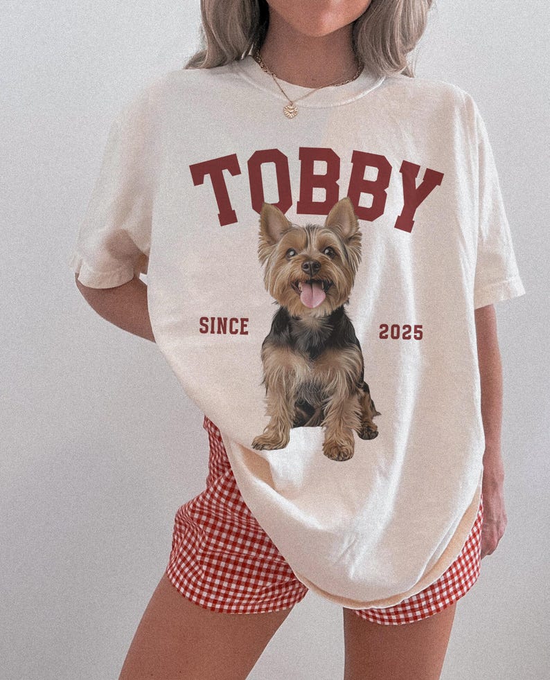 May include: Cream-coloured t-shirt featuring the name "TOBBY" in maroon block letters and a Yorkshire Terrier graphic. The text "SINCE 2025" is below the dog. The person is wearing red and white checkered shorts.
