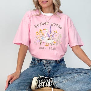 May include: Pink t-shirt with a white collar featuring a graphic of a goose surrounded by flowers and the text "Mother Goose est. 2025". The shirt is paired with blue jeans.