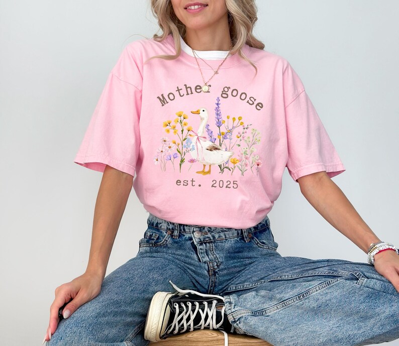 May include: Pink t-shirt with a white collar featuring a graphic of a goose surrounded by flowers and the text "Mother Goose est. 2025". The shirt is paired with blue jeans.