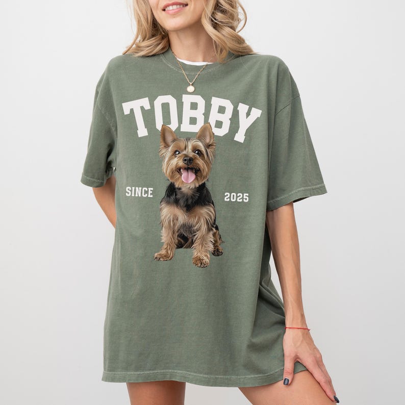 May include: Sage green t-shirt featuring the name "TOBBY" in white block letters and a Yorkshire Terrier dog graphic. The text "SINCE 2025" is below the dog. The shirt has a loose, oversized fit.