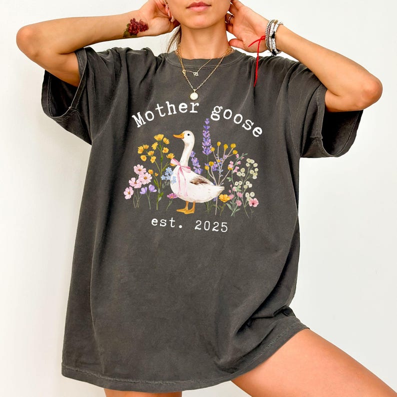 May include: Dark gray t-shirt featuring a whimsical design. The shirt displays the words "Mother Goose" above a charming illustration of a goose surrounded by colorful flowers. The text "est. 2025" is printed below the goose.