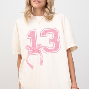 May include: A white t-shirt with a pink number "13" printed on the front. The number is in a varsity style font with a pink bow underneath.