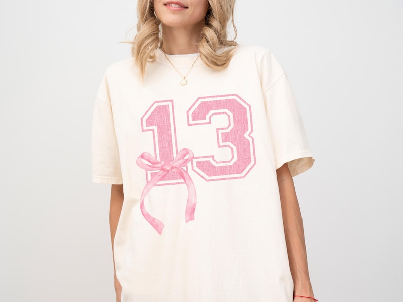 May include: A white t-shirt with a pink number "13" printed on the front. The number is in a varsity style font with a pink bow underneath.