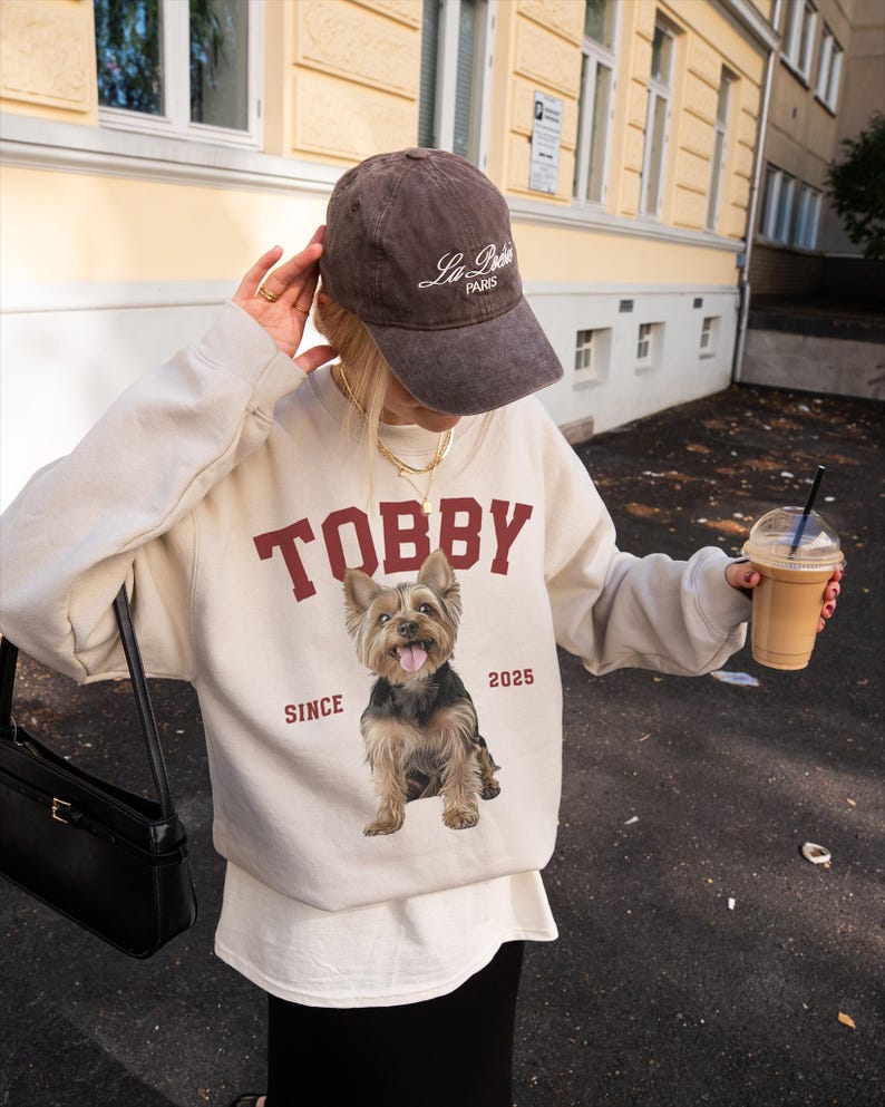 May include: A beige sweatshirt with the name "TOBBY" in maroon lettering and a Yorkshire Terrier graphic. The sweatshirt also features the text "SINCE 2025". The person is wearing a brown baseball cap with "La Paris" printed on it.