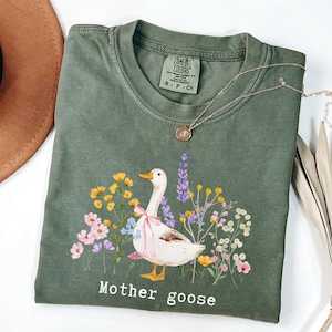 May include: An olive green t-shirt featuring a graphic of a white goose with a pink bow, surrounded by colorful flowers. The words "Mother goose" are printed below the illustration. A gold necklace and a brown hat are also visible.