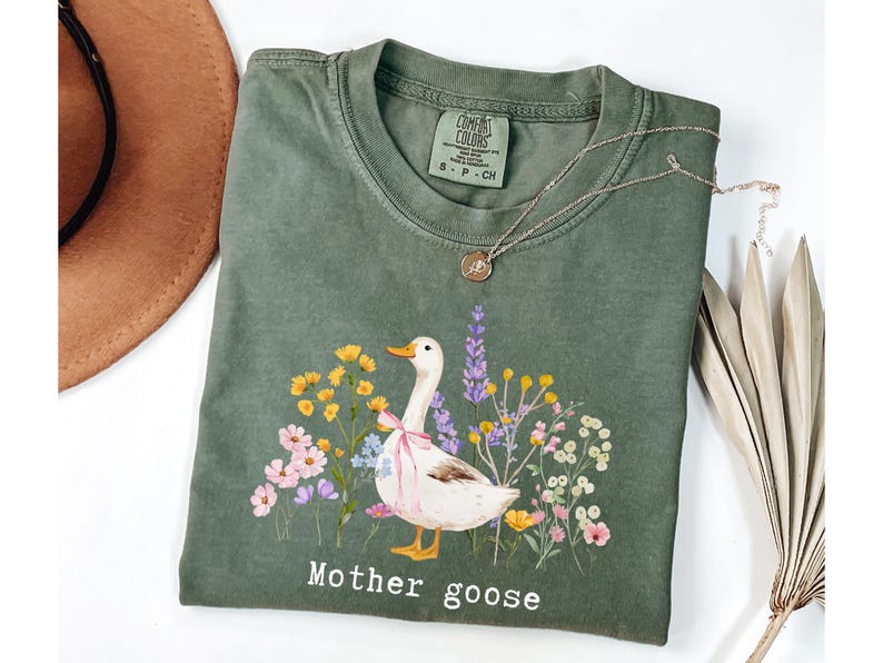 May include: An olive green t-shirt featuring a graphic of a white goose with a pink bow, surrounded by colorful flowers. The words "Mother goose" are printed below the illustration. A gold necklace and a brown hat are also visible.