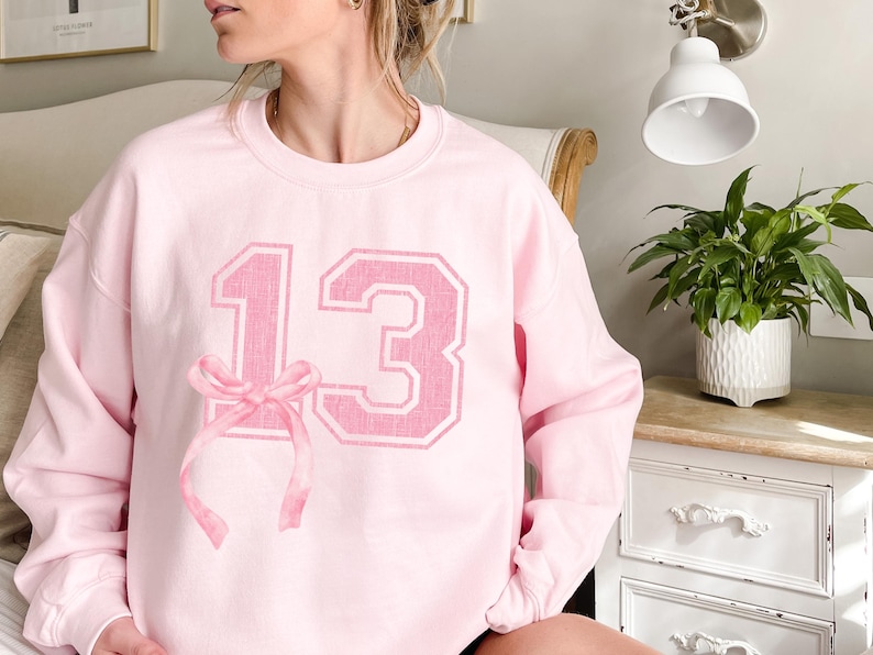 May include: Pink sweatshirt with a large pink number "13" printed on the front. A pink ribbon is tied around the number.