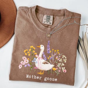 May include: A brown Comfort Colors t-shirt featuring a whimsical illustration of a goose with a pink bow, surrounded by colorful flowers. The text "Mother goose" is printed below the design. A silver necklace with a pendant is on the shirt.