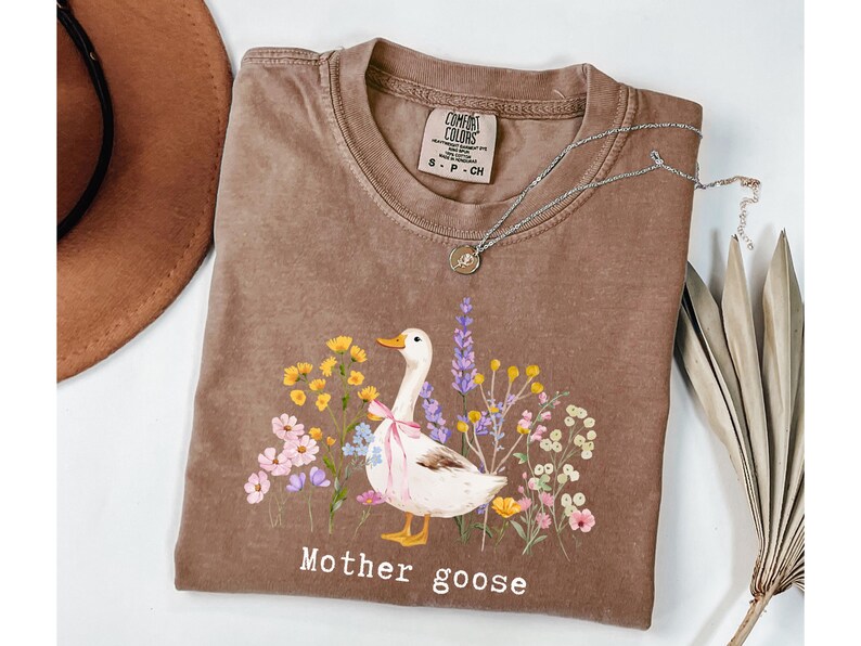 May include: A brown Comfort Colors t-shirt featuring a whimsical illustration of a goose with a pink bow, surrounded by colorful flowers. The text "Mother goose" is printed below the design. A silver necklace with a pendant is on the shirt.