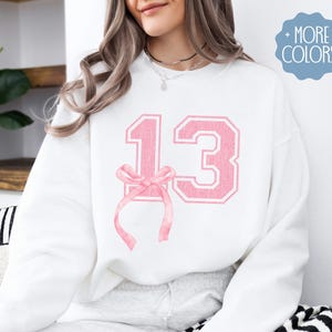 May include: White sweatshirt with a pink number 13 and a pink bow on the front.