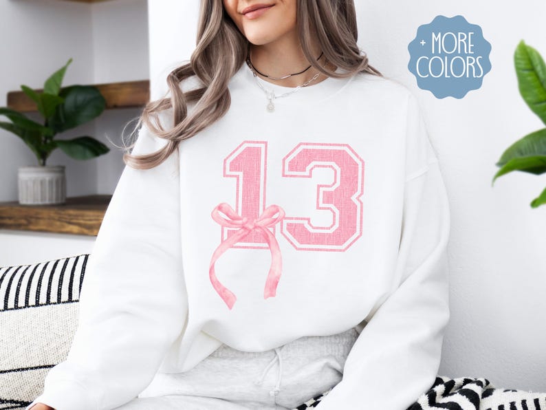 May include: White sweatshirt with a pink number 13 and a pink bow on the front.