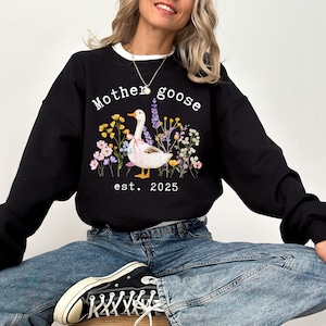 May include: Black sweatshirt with a white collar featuring a graphic of a goose with a pink bow, surrounded by flowers. The text "Mother goose" and "est. 2025" are printed above and below the goose illustration.
