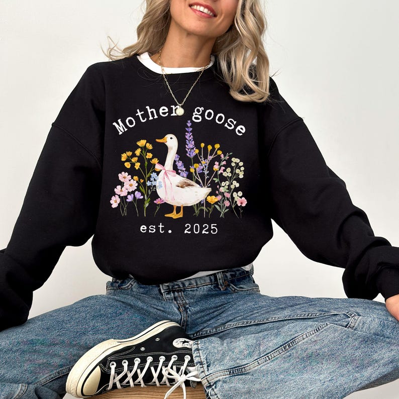 May include: Black sweatshirt with a white collar featuring a graphic of a goose with a pink bow, surrounded by flowers. The text "Mother goose" and "est. 2025" are printed above and below the goose illustration.