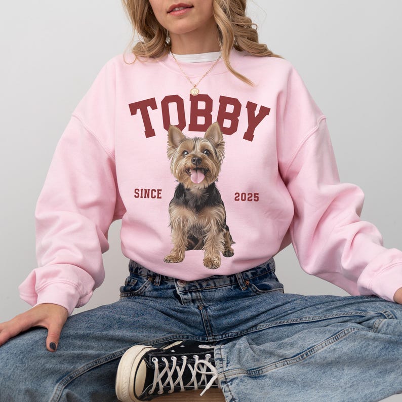 May include: A pale pink sweatshirt featuring the name "TOBBY" in maroon lettering. A Yorkshire Terrier dog graphic is centered below the name. The words "SINCE" and "2025" are printed on either side of the dog.
