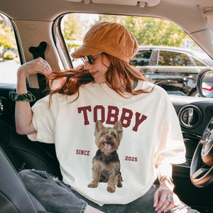 May include: Cream-coloured t-shirt featuring the name "TOBBY" in maroon block letters and a Yorkshire Terrier graphic. The text "SINCE 2025" is below the dog image. The person is wearing a brown cap and sunglasses.
