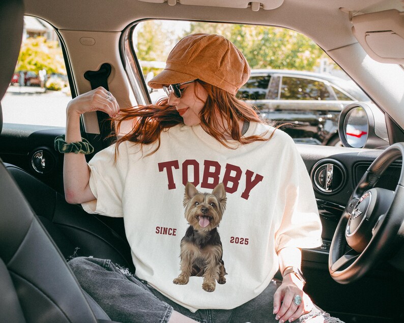 May include: Cream-coloured t-shirt featuring the name "TOBBY" in maroon block letters and a Yorkshire Terrier graphic. The text "SINCE 2025" is below the dog image. The person is wearing a brown cap and sunglasses.