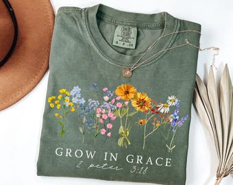May include: Olive green t-shirt with a floral design and the text "GROW IN GRACE 2 Peter 3:18". A silver necklace with a pendant rests on the shirt. The shirt is folded and next to a brown hat and dried plant.