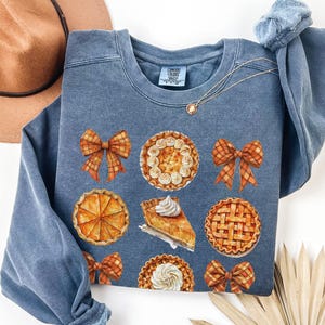 May include: A blue sweatshirt with a Thanksgiving theme. It features images of pumpkin pies, pecan pies, and bows in shades of orange and brown.