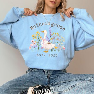 May include: Light blue sweatshirt with the text "Mother goose est. 2025" and a graphic of a goose wearing a bow, surrounded by colorful flowers. The sweatshirt is a long-sleeved crew neck.