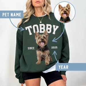 May include: A dark green sweatshirt featuring the name "TOBBY" in white block letters. A Yorkshire Terrier image is centered below the name. The words "SINCE" and "2025" are printed on either side of the dog image. A smaller dog photo is in the upper right corner.