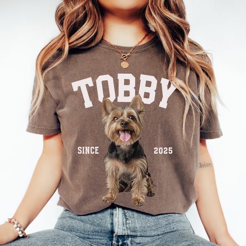 May include: Brown t-shirt featuring the name "TOBBY" in white block letters and a Yorkshire Terrier graphic. The text "SINCE 2025" is printed below the name. The dog is brown, black, and tan with its tongue out.
