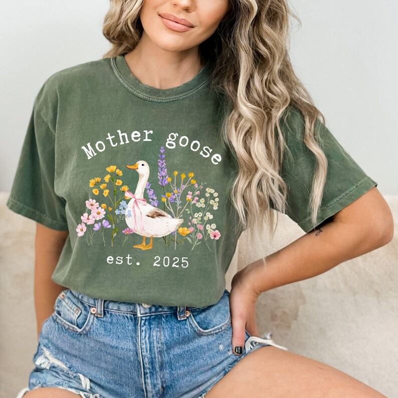 May include: Olive green t-shirt with the words "Mother Goose" above a watercolor illustration of a goose surrounded by flowers. The text "est. 2025" is below the illustration. The shirt is a comfortable, casual style.