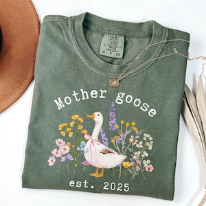 May include: Olive green t-shirt with the words "Mother Goose" and a watercolor illustration of a goose with a pink bow, surrounded by flowers. The text "est. 2025" is below the illustration. A gold necklace is on the shirt.