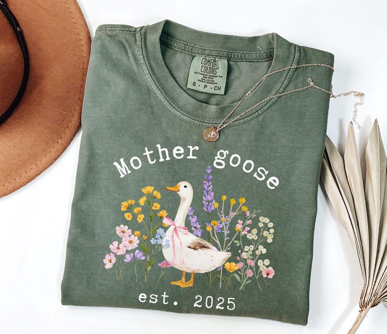 May include: Olive green t-shirt with the words "Mother Goose" and a watercolor illustration of a goose with a pink bow, surrounded by flowers. The text "est. 2025" is below the illustration. A gold necklace is on the shirt.