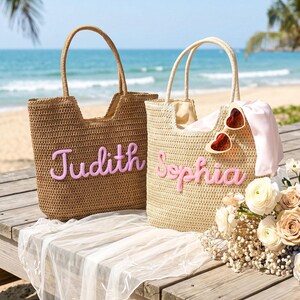 May include: Two woven straw tote bags with top handles. The left bag is brown with the name Judith in pink script. The right bag is beige with the name Sophia in pink script, featuring heart-shaped sunglasses and a light pink fabric draped inside.