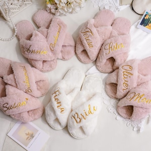 May include: A collection of plush, open-toe slippers with crisscross straps in soft pink and white. Each pair features gold embroidered names and titles including Anna, Bridesmaid, Teresa, Sister, Susan, Fiona, Bride, Oliver, and Mom.