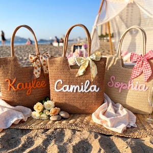 May include: Three woven straw tote bags with two handles each. The bags feature personalized names in raised script: Kaylee in orange, Camila in white, and Sophia in pink. Each bag is decorated with a patterned ribbon bow tied to one handle.
