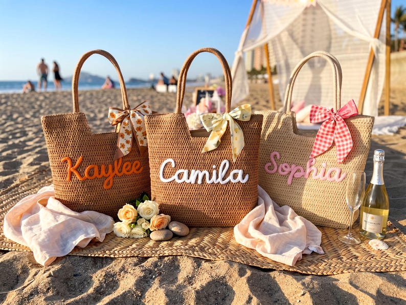 May include: Three woven straw tote bags with two handles each. The bags feature personalized names in raised script: Kaylee in orange, Camila in white, and Sophia in pink. Each bag is decorated with a patterned ribbon bow tied to one handle.