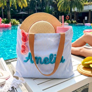 May include: A blue and white striped tote bag with the name "Amelia" embroidered in teal. The bag contains a straw hat, pink heart-shaped sunglasses, and a pink tumbler. The bag has brown straps and is sitting on a white table near a pool.