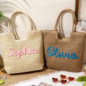 May include: Two woven straw tote bags with dual handles. The left bag is light beige with the name Sophia in raised pink script. The right bag is tan with the name Olivia in raised blue script. Both bags feature a rectangular shape with an open top.