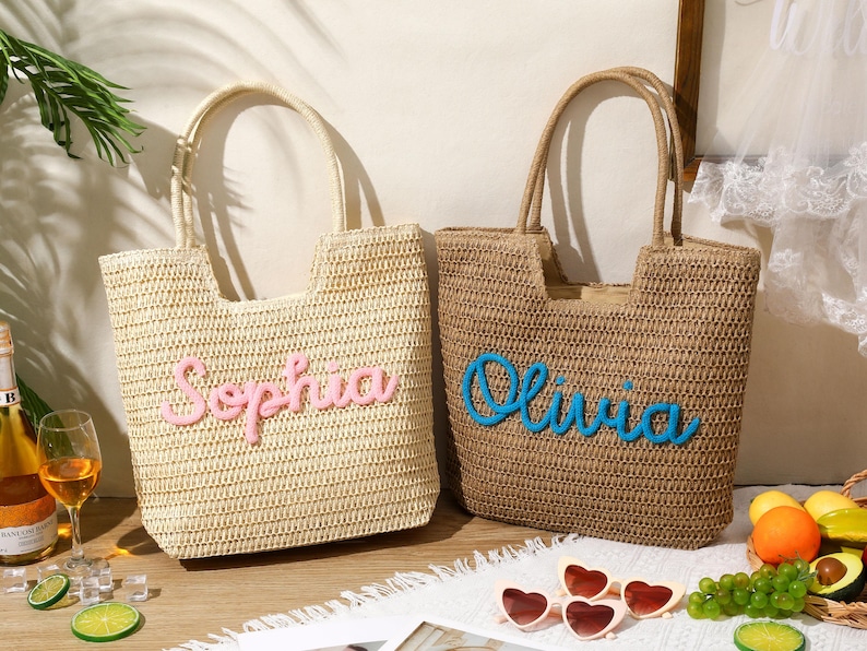 May include: Two woven straw tote bags with dual handles. The left bag is light beige with the name Sophia in raised pink script. The right bag is tan with the name Olivia in raised blue script. Both bags feature a rectangular shape with an open top.