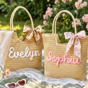 May include: Two woven straw tote bags with dual handles. The left bag features the white embroidered name Evelyn and a brown floral ribbon. The right bag features the pink embroidered name Sophia and a pink floral ribbon, both with open tops.