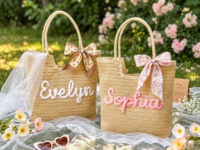 May include: Two woven straw tote bags with dual handles. The left bag features the white embroidered name Evelyn and a brown floral ribbon. The right bag features the pink embroidered name Sophia and a pink floral ribbon, both with open tops.
