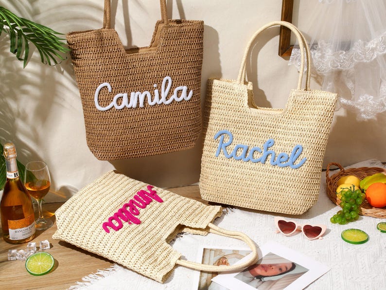 May include: Three woven straw tote bags with two top handles each. One tan bag features the white embroidered name Camila. One light beige bag features the blue embroidered name Rachel. One light beige bag features the pink embroidered name Amelia.