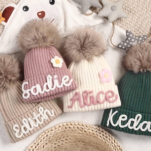 Peut inclure: Quatre bonnets en tricot avec des pompons en fausse fourrure et des noms personnalisés. Les bonnets sont rose, beige, blanc et vert. Les noms sont Sadie, Edith, Alice et Kedar.