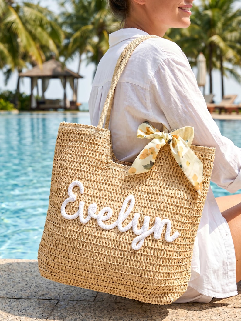 May include: A tan woven straw tote bag featuring the name Evelyn in white cursive rope lettering on the front. The bag includes two shoulder straps and a decorative light yellow fabric ribbon tied to one side with a subtle lemon print pattern.