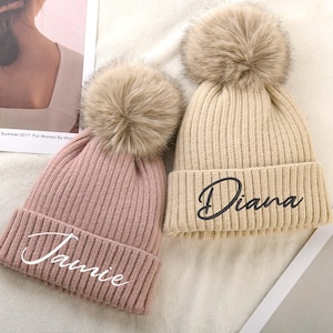May include: Two knitted beanies with faux fur pom poms. One beanie is pink with the name "Jamie" embroidered in white. The other beanie is beige with the name "Diana" embroidered in black.
