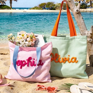 Puede incluir: Dos bolsas de playa en una playa de arena. Una es rosa con correas azules y el nombre "Sophia" en rosa. La otra es verde con correas naranjas y el nombre "Olivia" en naranja. La escena de la playa incluye gafas de sol y flores.