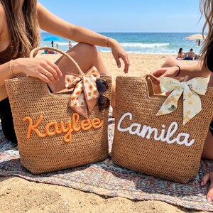 May include: Two woven straw tote bags featuring embroidered names. The left bag displays the name Kaylee in orange thread with a floral scarf, while the right bag shows the name Camila in white thread with a lemon-patterned scarf attached to the handle.